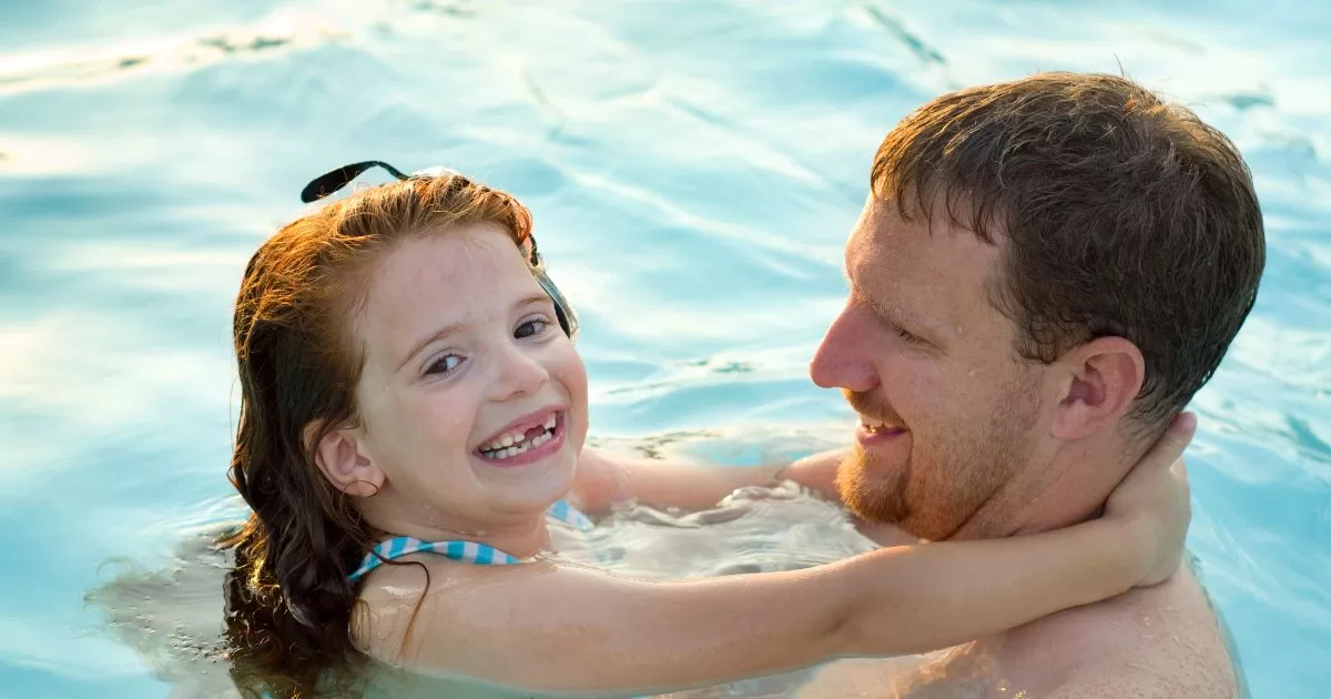 A father holds his daughter in the pool. They are both smiling and having fun.